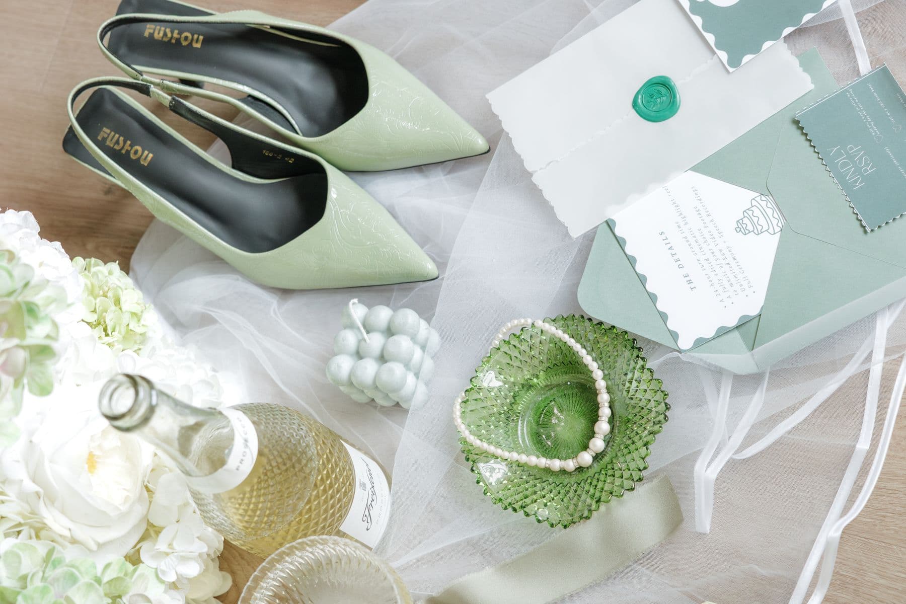 A sage-green flat lay of heels, wax-sealed stationery, pearls and a glass dish on tulle